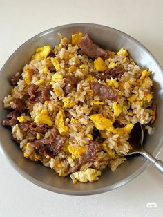 a bowl filled with rice and meat on top of a white table next to a spoon