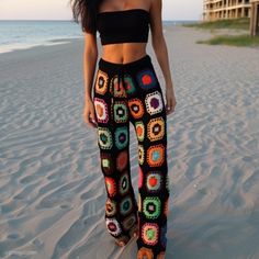 a woman standing on top of a beach next to the ocean wearing colorful crocheted pants