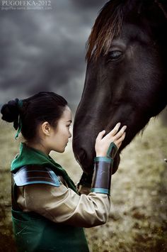 a woman is petting the nose of a horse in a field with dark clouds behind her