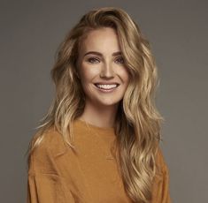 a woman with long blonde hair wearing a brown sweater and black pants, smiling at the camera