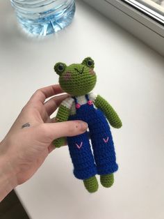 a hand holding a crocheted frog toy in front of a window