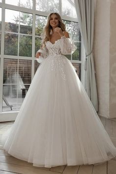 a woman standing in front of a window wearing a white wedding dress with sheer sleeves