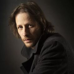 a man with long hair wearing a black coat and looking at the camera while standing in front of a dark background