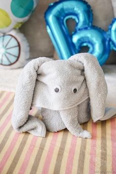 an elephant stuffed animal sitting on top of a striped table cloth next to blue balloons