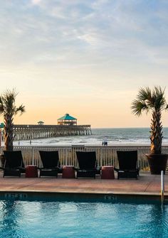 lounge chairs sitting on the edge of a swimming pool next to an ocean with a pier in the background