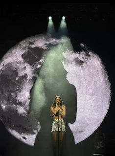 a woman standing on stage in front of a giant moon with her hands behind her head