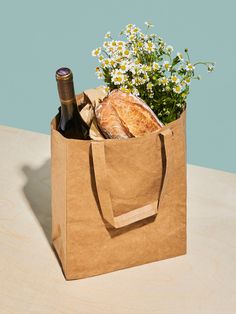 a brown paper bag with bread and flowers in it sitting on a table next to a bottle of wine