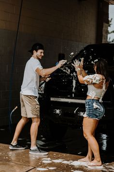 a man and woman are washing their hands in front of a black truck with foam on it