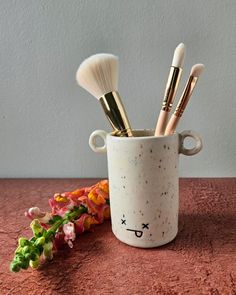 a cup filled with makeup brushes sitting on top of a table next to some flowers