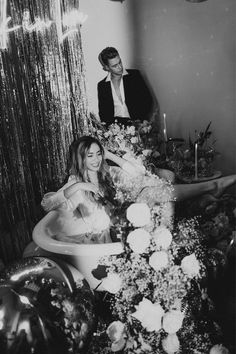 black and white photograph of woman in bathtub surrounded by flowers with man standing next to her