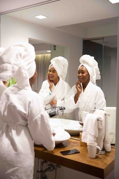 three women in white robes are brushing their teeth and looking at each other through the mirror