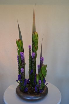 an arrangement of purple flowers and greenery in a vase on a white table top