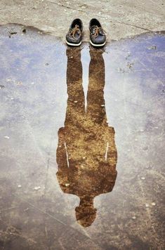 a pair of shoes sitting on top of a puddle next to a sidewalk with the reflection of someone's feet in it