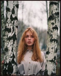 a woman standing between two tall trees in the middle of a forest, looking at the camera