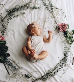 a baby is laying on a white blanket surrounded by flowers and greenery with her hands in the air