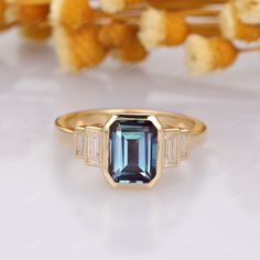 an emerald and baguette cut diamond ring on a white surface with dried flowers in the background