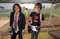 two young men are playing guitars under an awning in front of a fenced area