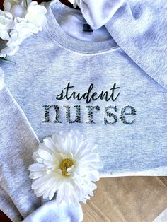 a white flower sitting on top of a wooden table next to a gray sweatshirt that says student nurse