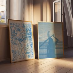 two framed pictures sitting on top of a wooden floor next to a window with curtains