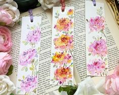 three bookmarks with watercolor flowers on them sitting next to an open book and some books