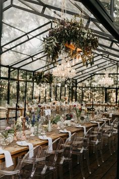 a long table with clear chairs under a chandelier filled with flowers and greenery