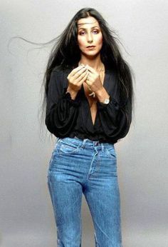 a woman with long black hair standing in front of a gray background wearing jeans and heels