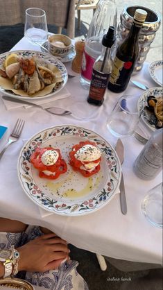 a table topped with plates of food next to bottles of wine and silverware on top of a white cloth covered table