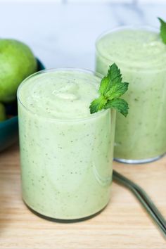 two glasses filled with green smoothie on top of a wooden table next to fruit