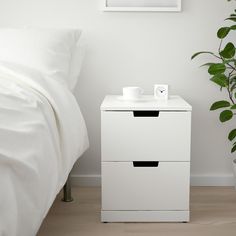 a white night stand next to a bed with a plant on it and a window in the background