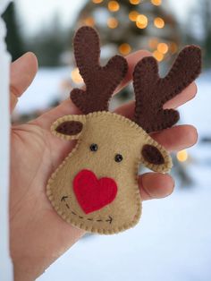 a hand holding a small felt reindeer ornament in front of a christmas tree