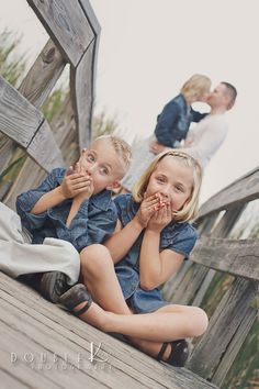 two young children sitting on a wooden bridge with their hands in their mouths and one child standing behind them