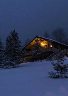 a cabin lit up at night with snow on the ground and pine trees in front