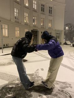 two people standing in the snow touching each other's hands with buildings in the background