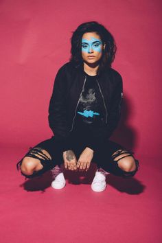 a woman with blue makeup sitting on the floor in front of a pink background wearing white sneakers