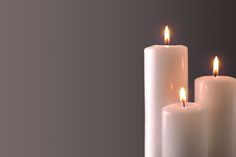 three white candles with one lit and the other turned on, in front of a dark background