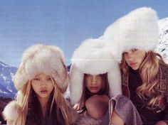 three women wearing fur hats and sitting on the ground in front of snow covered mountains