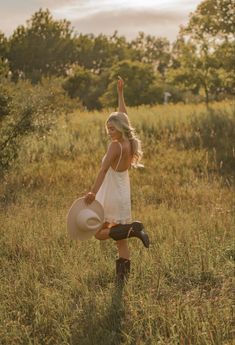 a woman in a white dress and cowboy boots standing in tall grass with her hat raised