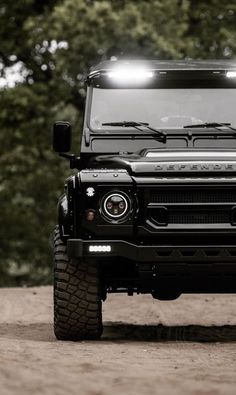 the front end of a black land rover with its lights on, parked in dirt area next to trees