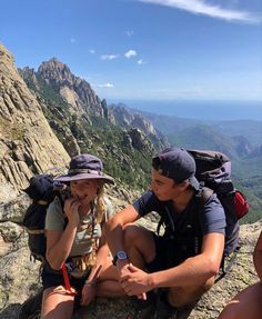 two people sitting on the side of a mountain talking on their cell phones and looking at each other