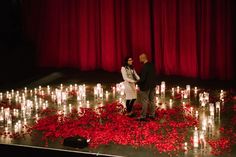 two people standing on a stage surrounded by candles and rose petals in front of a red curtain