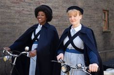 two women standing next to each other with bicycles in front of a brick wall and building