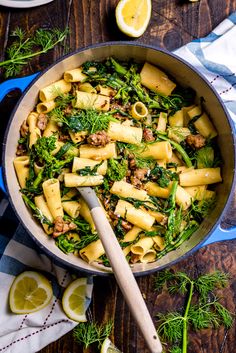 a skillet filled with pasta and vegetables on top of a wooden table next to sliced lemons