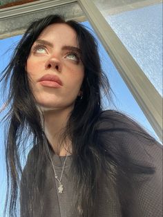 a woman with long black hair standing in front of a window looking up at the sky