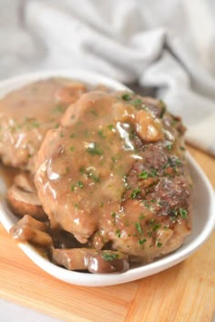 a white bowl filled with meat covered in gravy and mushrooms on top of a wooden cutting board