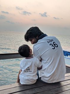 a man sitting next to a little boy on top of a wooden bench near the ocean