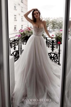 a woman standing in front of a window wearing a wedding dress and holding her hand on her head