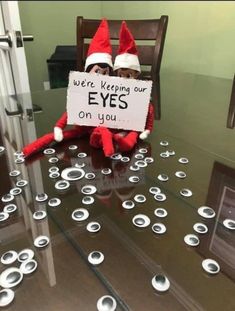 an elf sitting on top of a glass table holding a sign that says, we're keeping our eyes on you