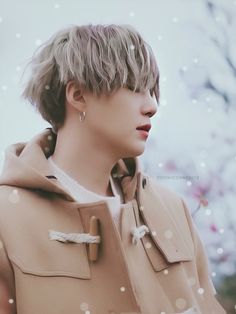 a man with short hair wearing a trench coat in front of snow covered trees and flowers