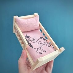 a hand holding a wooden baby crib with a pink blanket and horse drawn on it