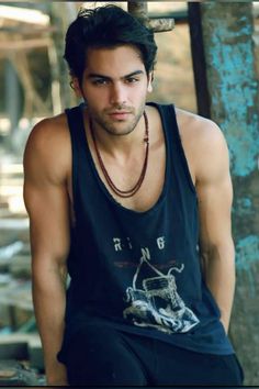 a young man wearing a black tank top sitting on a wooden bench next to a tree